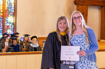 Two women stand together inside a church during a graduation ceremony. One wears academic regalia, while the other holds a certificate and a black gift bag. Graduates in caps and gowns are seated in the background, with stained-glass windows and wooden architectural details visible.